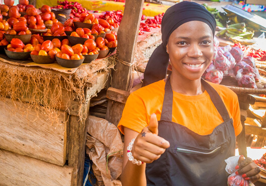 A woman wearing an orange t-shirt and black apron smiles and gives a thumbs up to the camera.