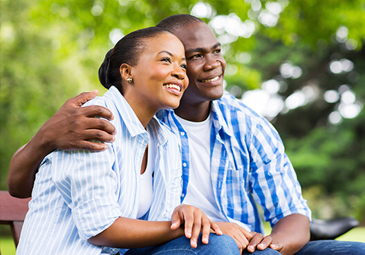 A couple wearing blue and white patterned shirts hug as they sit together on a bench in a park.